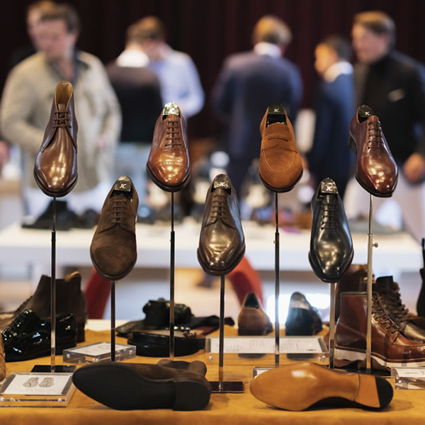 Diverse collectie herenschoenen uitgestald op een tafel tijdens een beurs, met mensen op de achtergrond. Elegant en overzichtelijk.