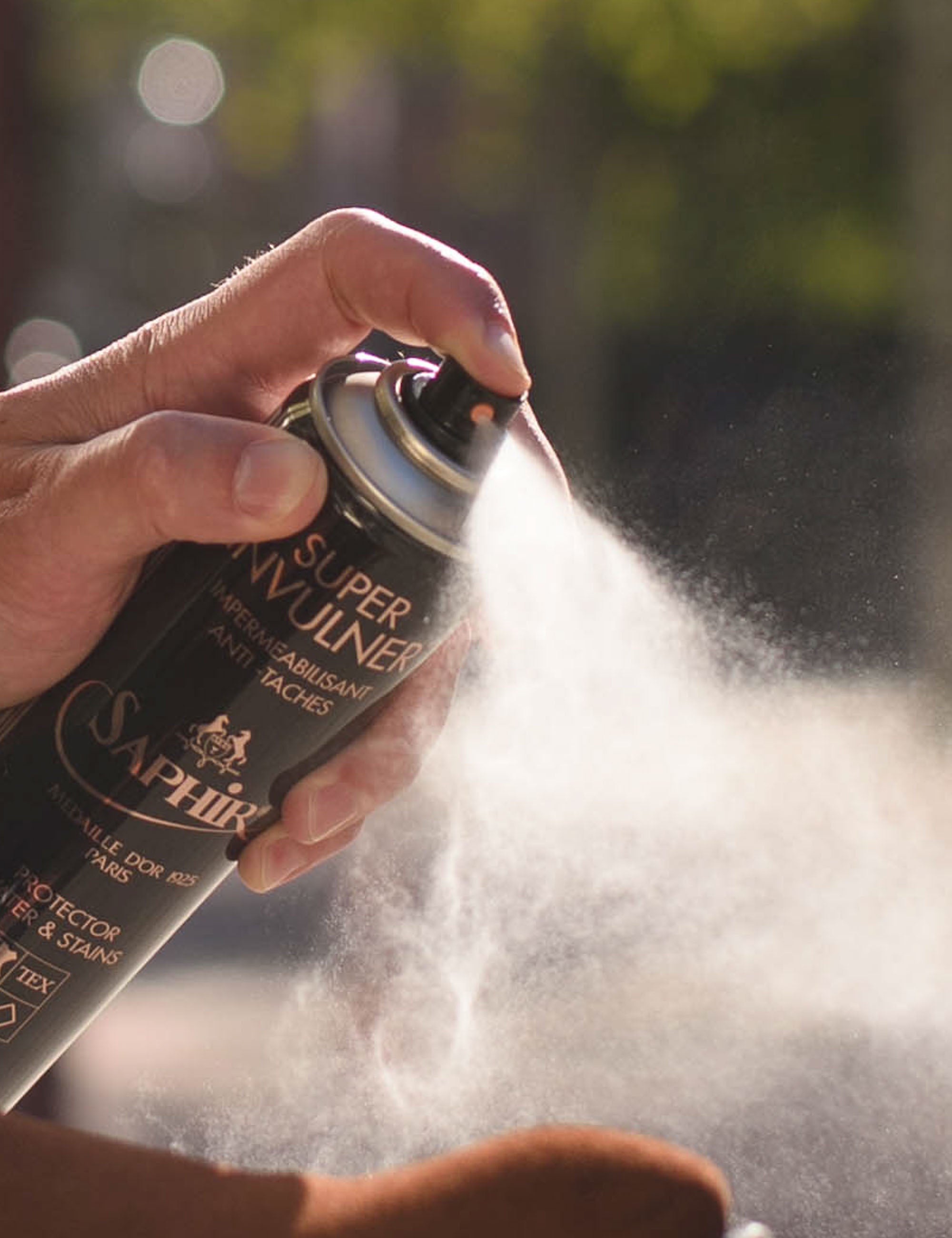 Een hand spuit Saphir Super Invulner schoenspray. De spray wolk is helder wit, de achtergrond is onscherp en groen. De tekst op de bus is leesbaar.