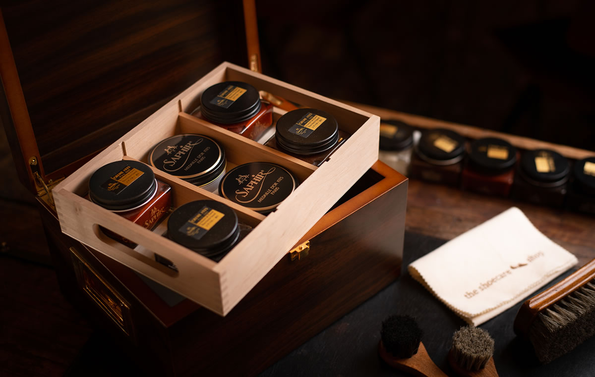 Houten schoenpoetsdoos met Saphir producten, borstels en doekje met "the shoecare-shop" tekst, sfeervolle belichting.