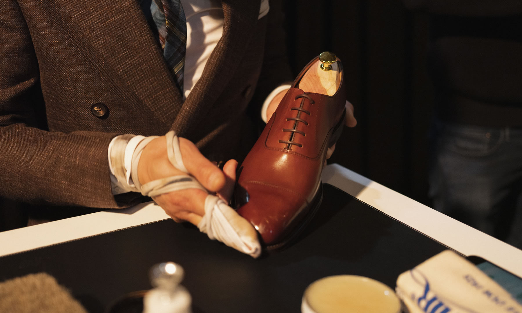Een man met een bruin jasje poetst een bruine leren schoen met een doek om zijn hand. Op tafel staat schoenpoets.