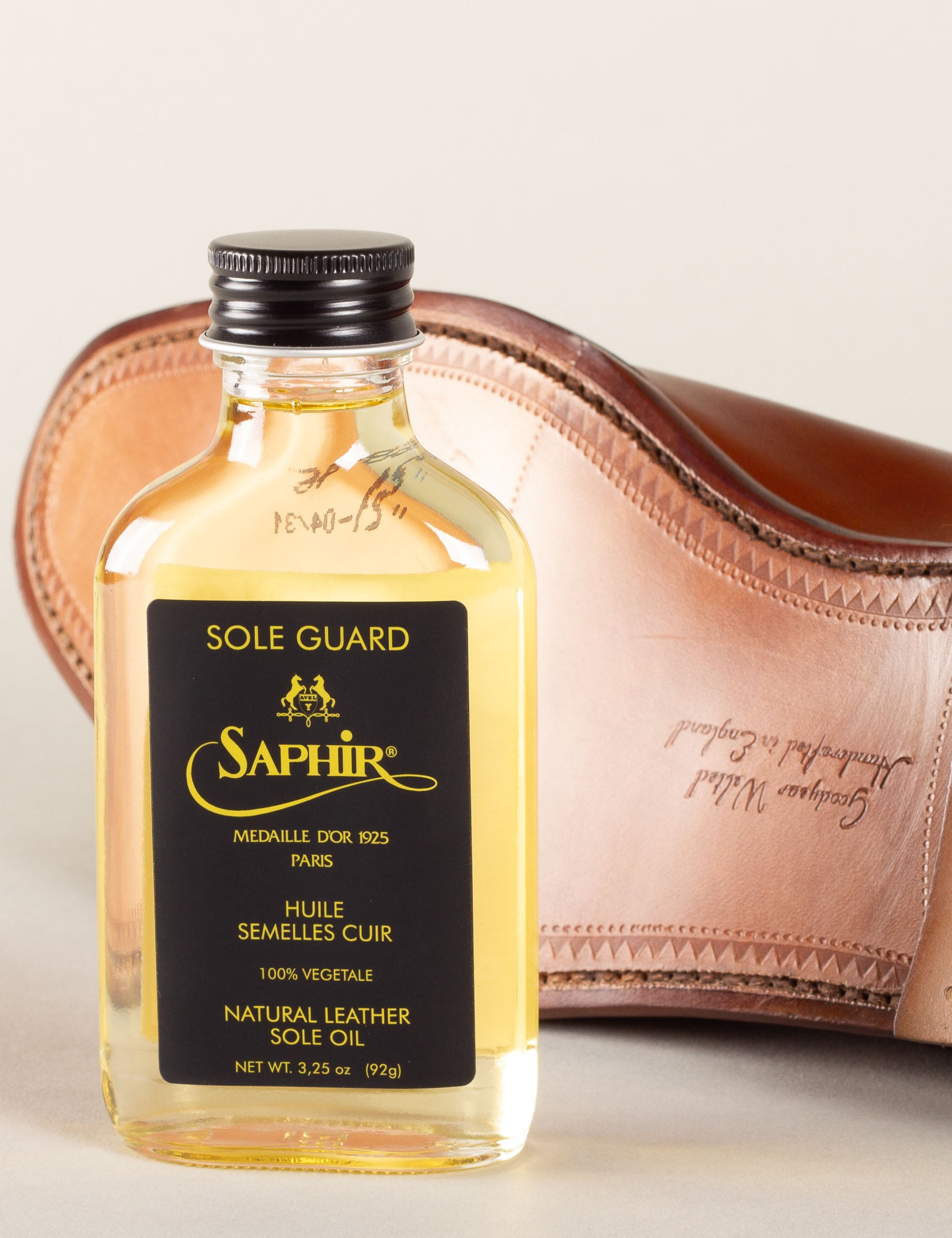 Flesje Saphir Sole Guard leerolie staat voor een lederen schoenzool met de tekst "Goodyear Welted Handcrafted in England". Goudgele olie tegen een crèmekleurige achtergrond.