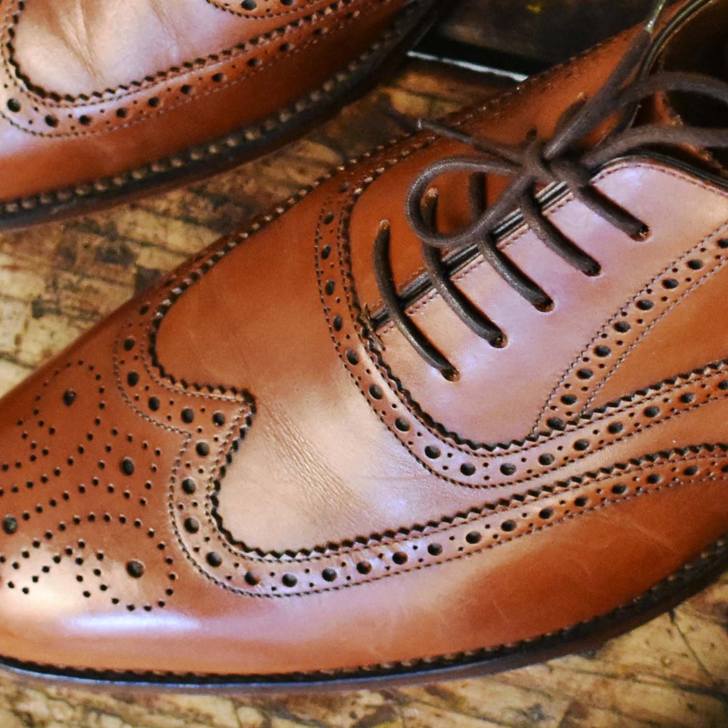 Close-up van een paar bruine leren brogues met veters op een houten oppervlak, met decoratieve perforaties en stiksels.