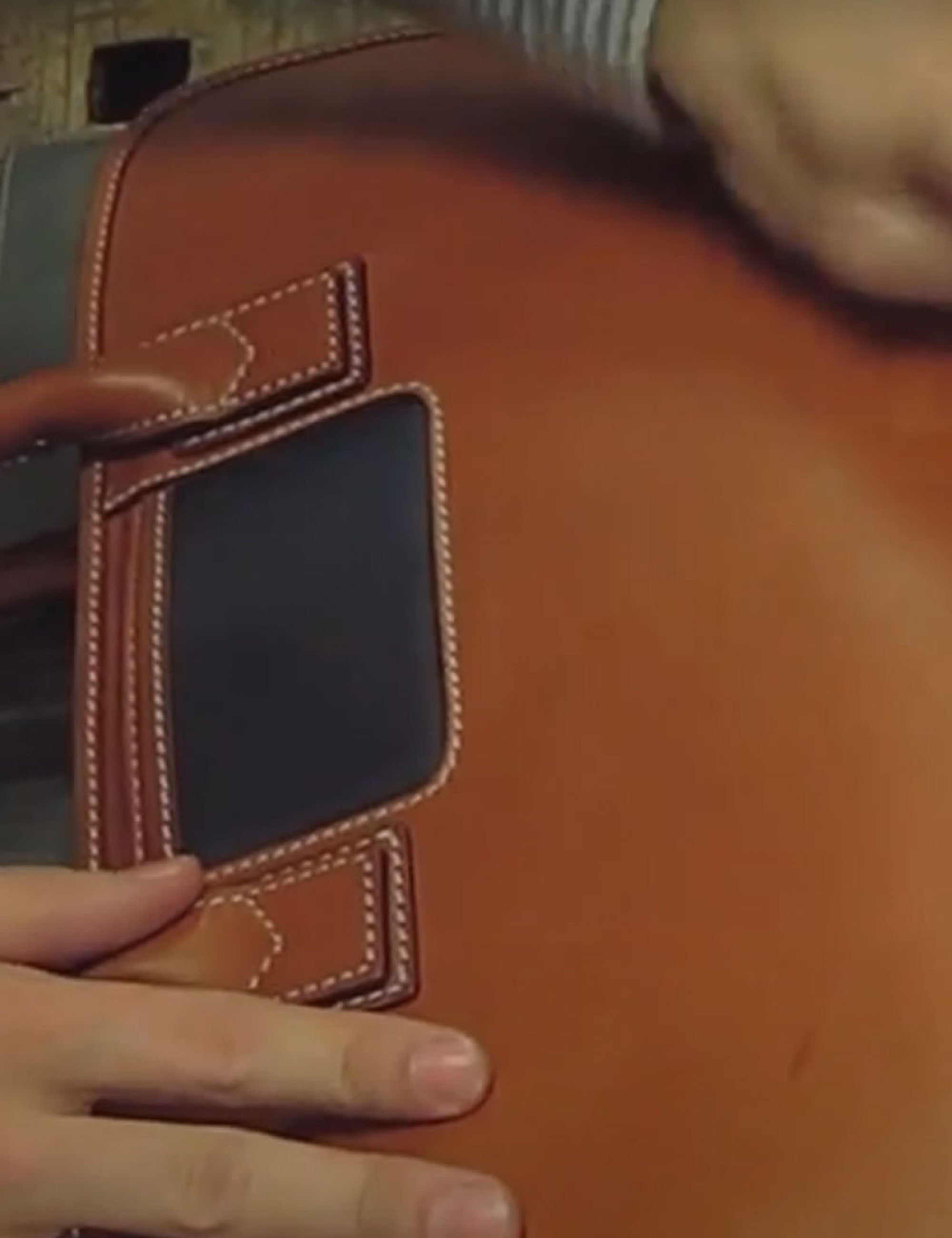 Een close-up van een bruine lederen tas met donkerblauwe accenten, witte stiksels, vastgehouden door twee handen.