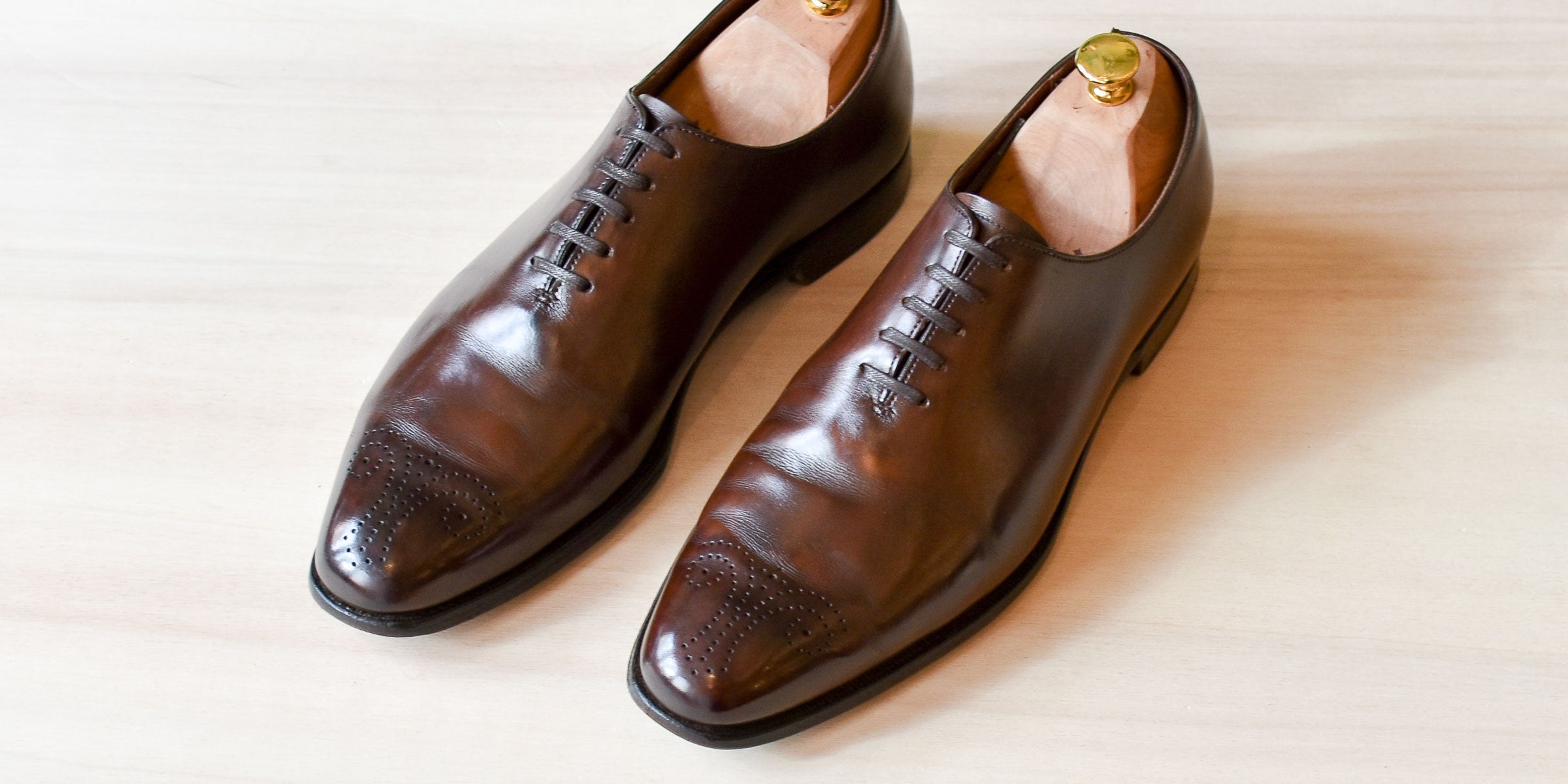 Twee glanzende, bruine herenschoenen met houten schoenspanners staan schuin op een lichte houten ondergrond.