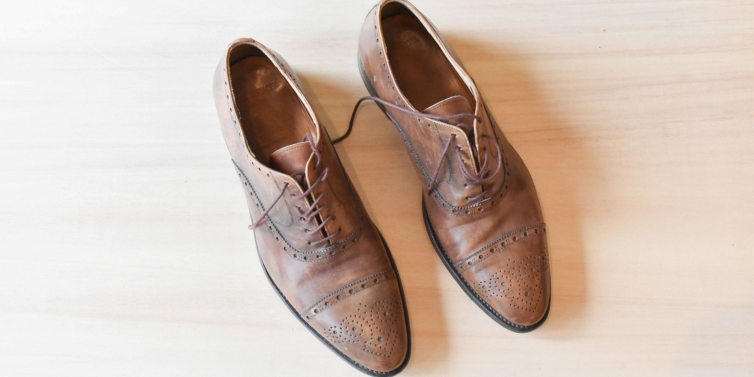 Twee bruine lederen herenschoenen met veters op een lichtbeige houten oppervlak, van bovenaf gefotografeerd.
