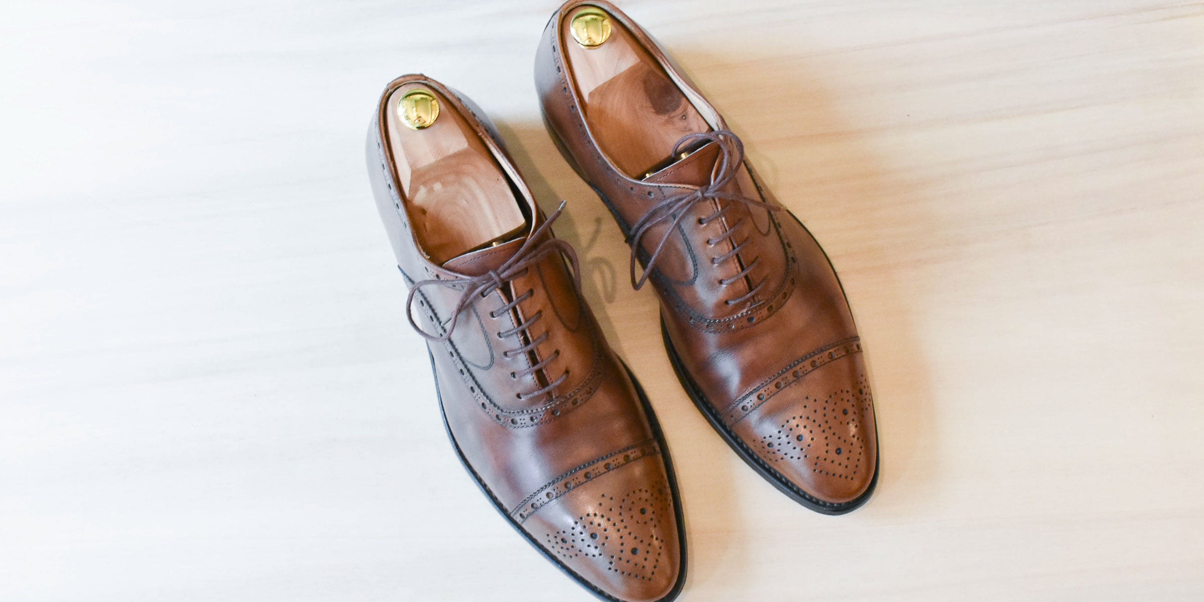 Een paar bruine leren herenschoenen met brogue details en houten schoenspanners op een witte houten ondergrond.