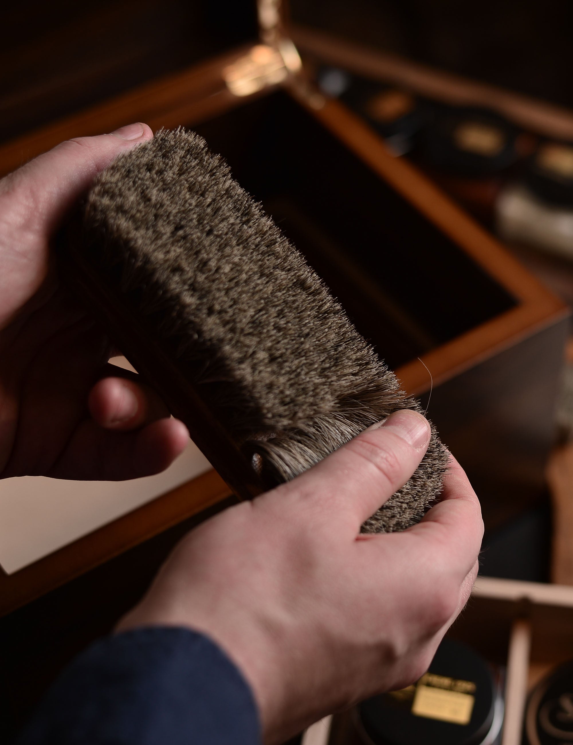 Twee handen houden een bruine borstel vast voor een open houten kist met zwarte potten. Warm licht.
