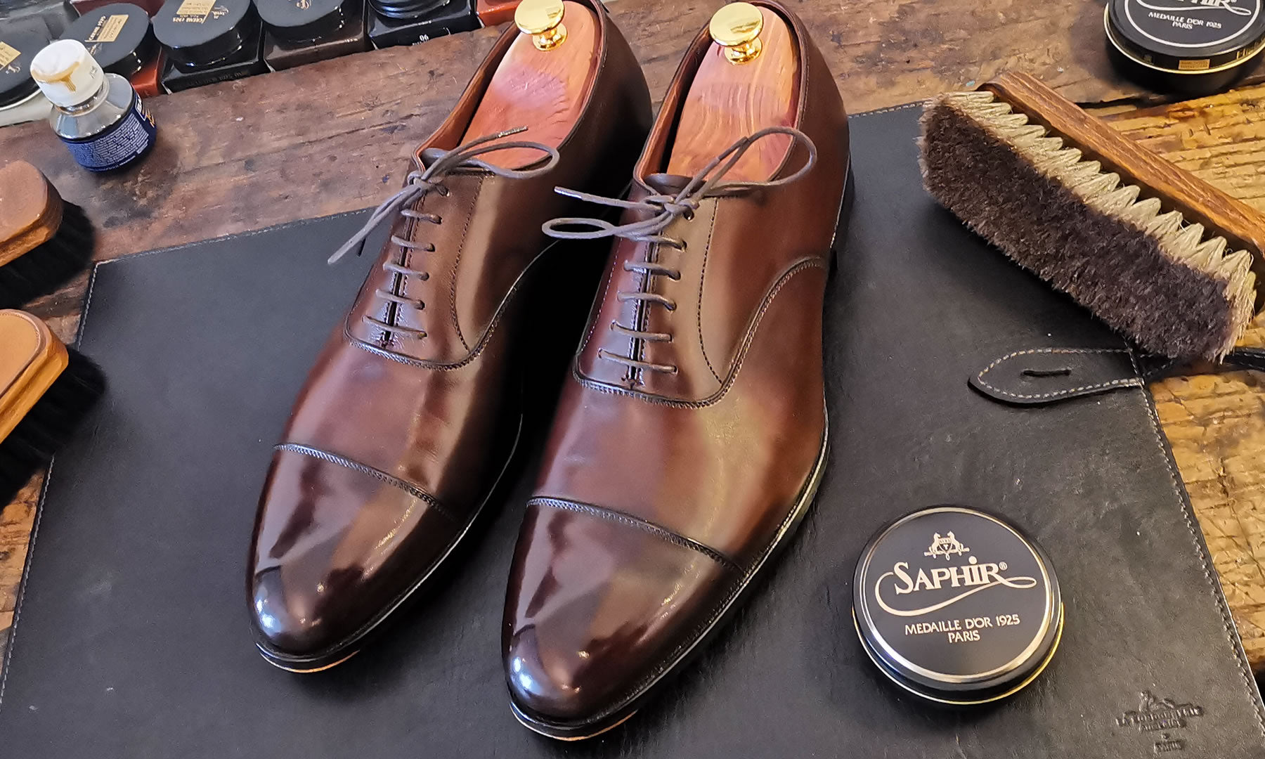 Twee gepoetste bruine herenschoenen met houten schoenspanners op een poetsmat, met Saphir schoenpoets en een borstel.