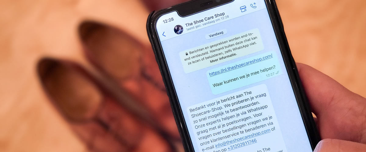 Een close-up van een smartphone in een hand, met een WhatsApp-gesprek van "The Shoe Care Shop" op het scherm, bruine schoenen op de achtergrond.